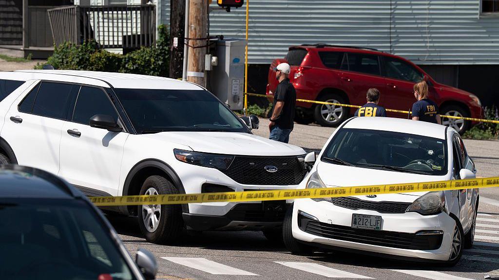 Dos coches blancos que colisionaron est&aacute;n detr&aacute;s de cinta policial amarilla. El parabrisas de uno de los coches tiene agujeros de bala.