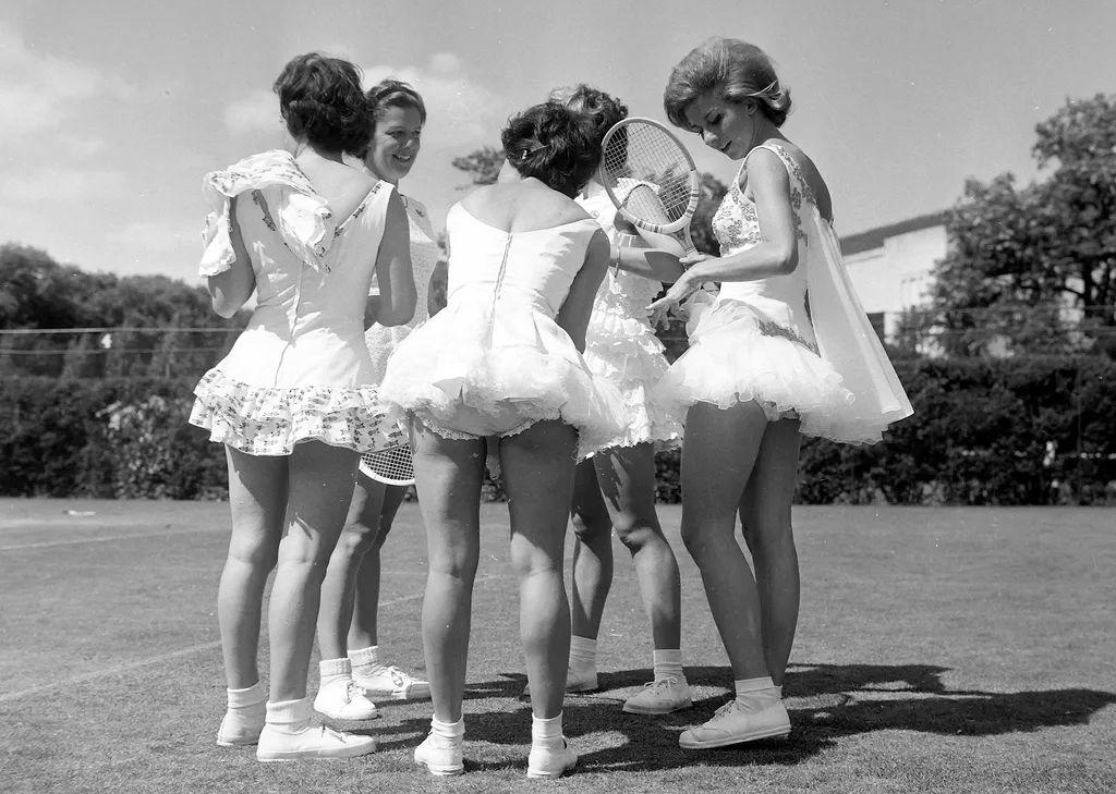 Fotograf&iacute;a en blanco y negro de cinco chicas conversando en c&iacute;rculo, todas con vestidos de tenis, mayoritariamente blancos y cortos, con volantes y encajes, mangas cortas. Todas calzan tenis y medias dobladas, sus piernas se ven casi completas. Se ven algunas raquetas. Est&aacute;n en una cancha de pasto y en el fondo se ve vegetaci&oacute;n. 