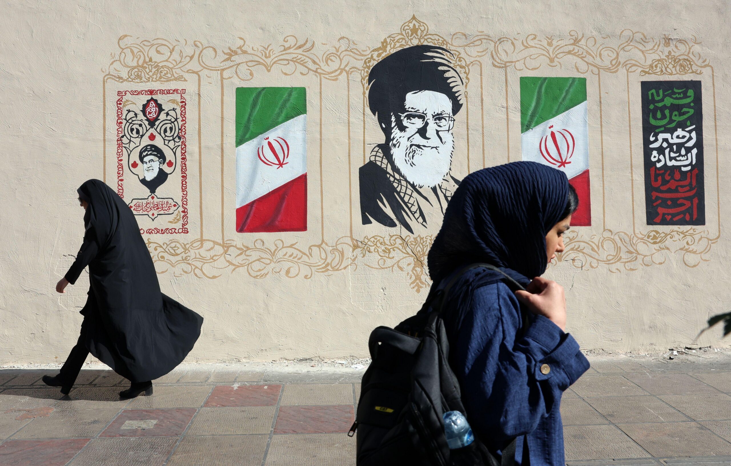 Dos mujeres con velo caminan por la acera frente a un mural que tiene banderas iran&iacute;es, caligraf&iacute;a decorativa y un gran retrato de Al&iacute; Jamenei. Una mujer lleva una t&uacute;nica negra, mientras que la otra viste un abrigo azul oscuro y lleva una mochila.