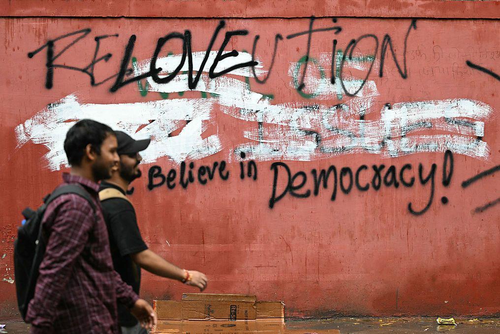 Simpatizantes del Partido Popular Cucaracha CJP) llegando a Jantar Mantar en Delhi caminan frente a un muro con grafiti que lee: “Revolución. Cree en la democracia”.