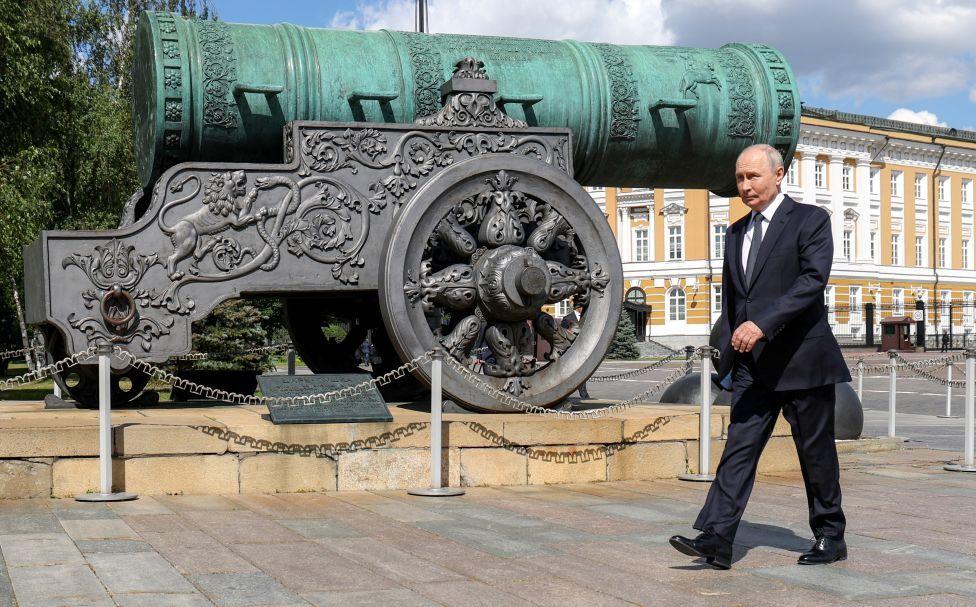 Putin pasa junto al monumento del Cañón del Zar en Moscú. Viste un traje oscuro, corbata azul y camisa blanca. El cañón, muy ornamentado, se alza sobre un pedestal.