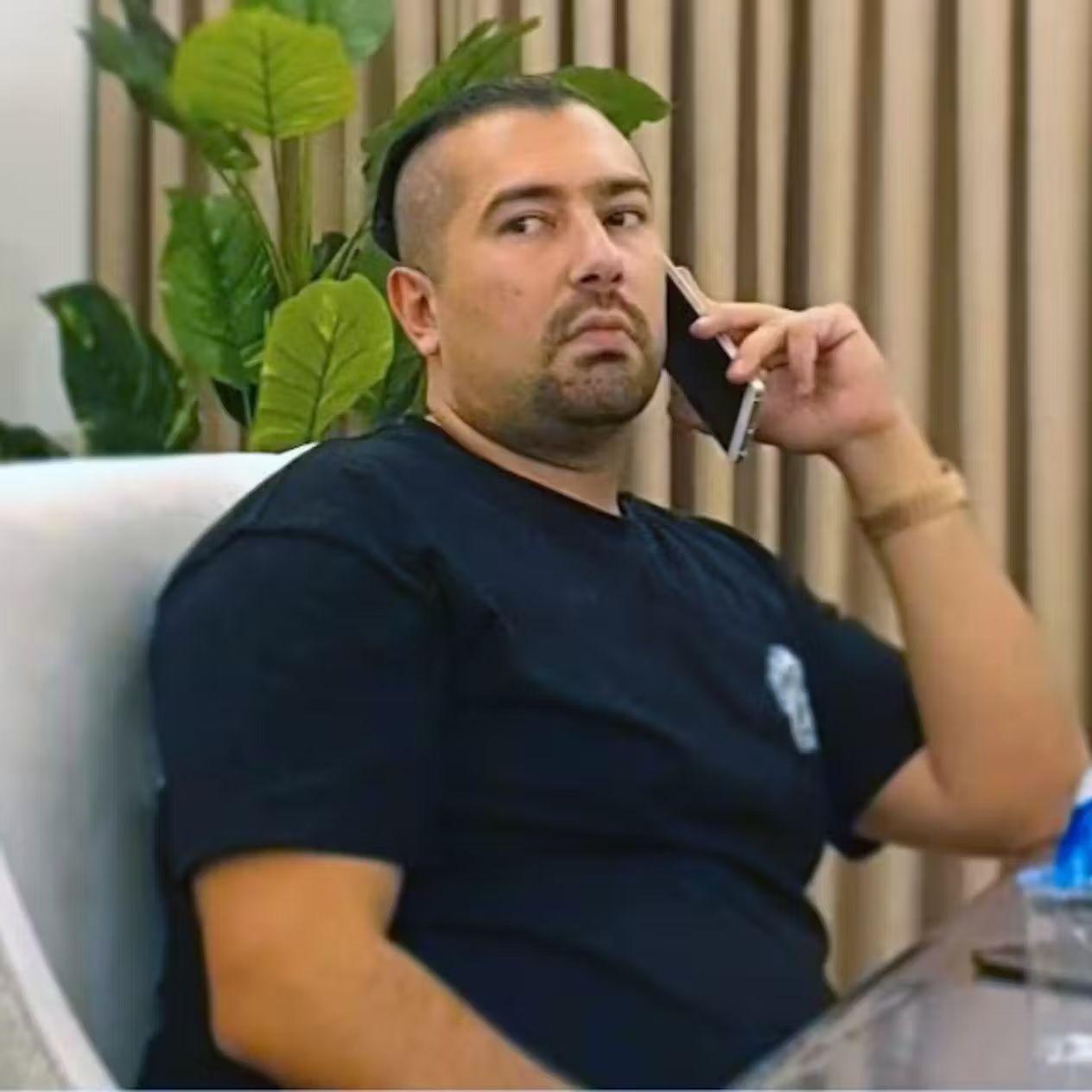 Rawa Majid habla por teléfono sentado en una silla. Lleva el pelo peinado hacia atrás y rapado a los lados, y luce una perilla. Mira hacia un lado con expresión concentrada.