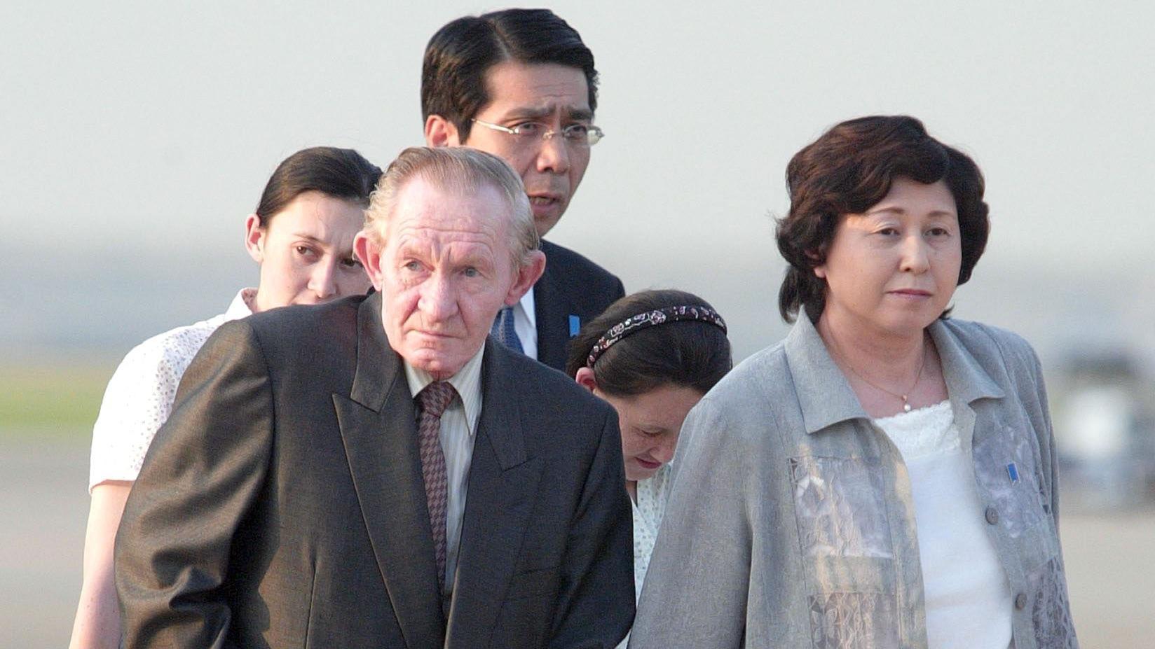 Charles Jenkins camina por la pista del aeropuerto, de la mano de Hitomi. Viste un traje gris oscuro. Hitomi también lleva un traje gris. Detrás de él están sus hijas, que visten camisas blancas (una de ellas mira hacia abajo), y un hombre japonés con traje.