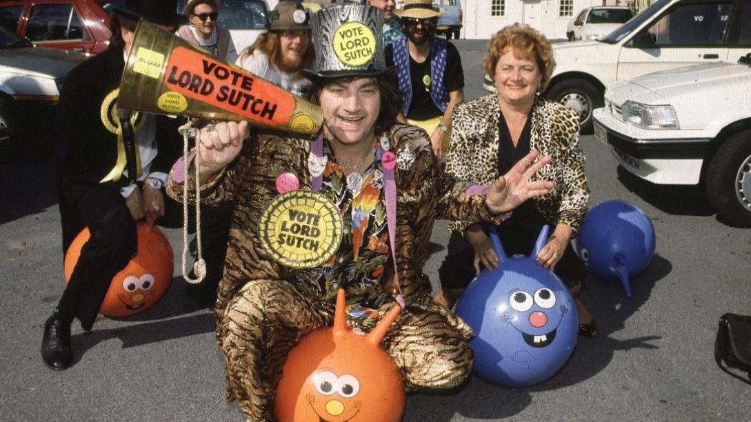 Lord Sutch, del Partido de los Locos, en su congreso de 1990. Viste un traje de piel de leopardo con un sombrero de copa a juego y está sentado sobre una pelota naranja. 