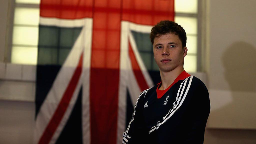 El atleta olímpico Freddie Woodward con los brazos cruzados y una gran bandera británica detrás.