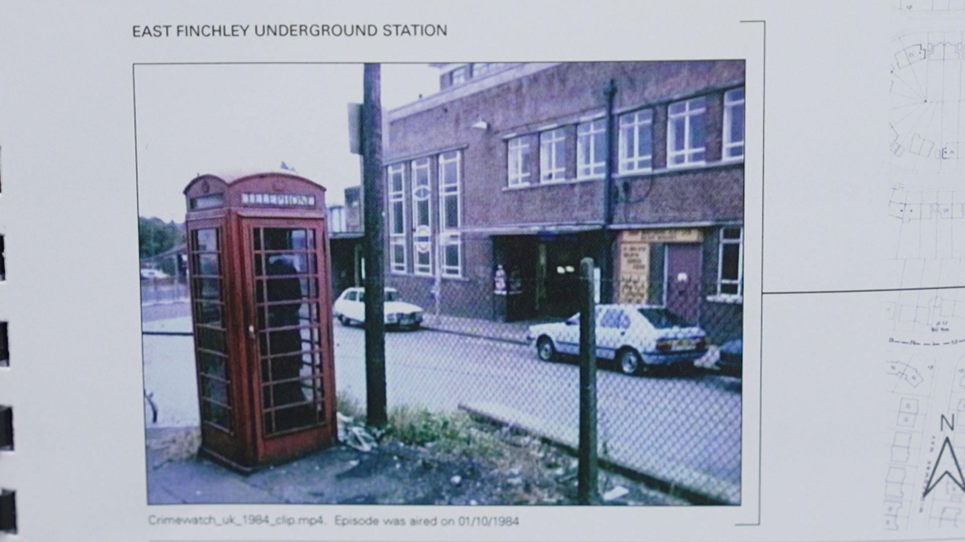 Fotografía en color de 1984 de una cabina telefónica roja tradicional situada en una esquina, con coches aparcados y edificios bajos detrás.