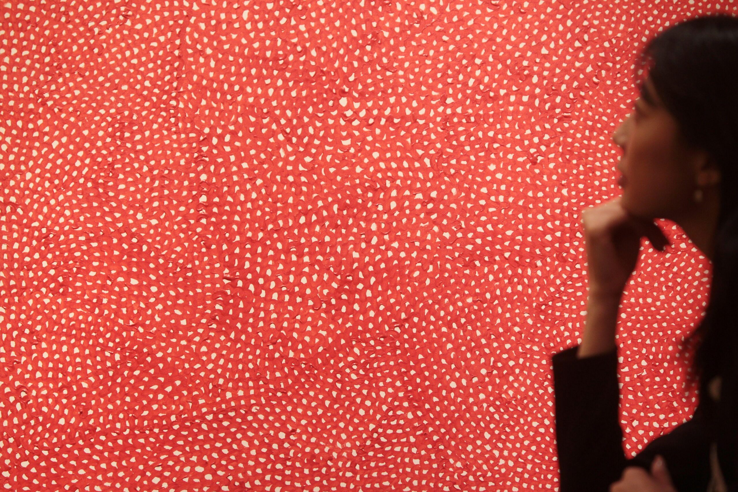 Una obra de arte con un estampado rojo y blanco llena la foto, con una mujer mirándola en el borde.