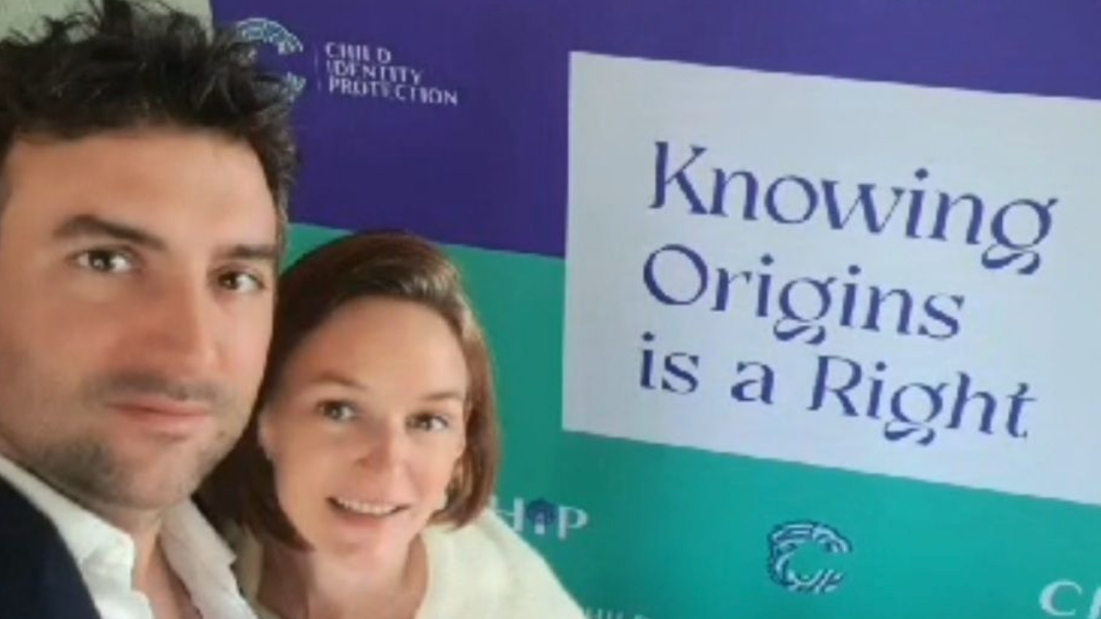 Audrey y Arthur posan juntos frente a una gran pancarta de campaña que muestra el mensaje "Conocer los orígenes es un derecho" e incluye logotipos asociados con la defensa de la identidad infantil.