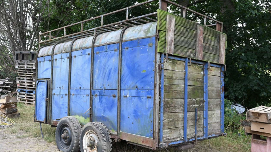 Un remolque cerrado de gran tamaño se encuentra sobre una superficie de hierba seca y tierra en un patio al aire libre, rodeado de árboles y diversos materiales almacenados. El remolque está pintado principalmente de azul y presenta un aspecto envejecido, con signos visibles de óxido, suciedad y desgaste en la estructura metálica. 