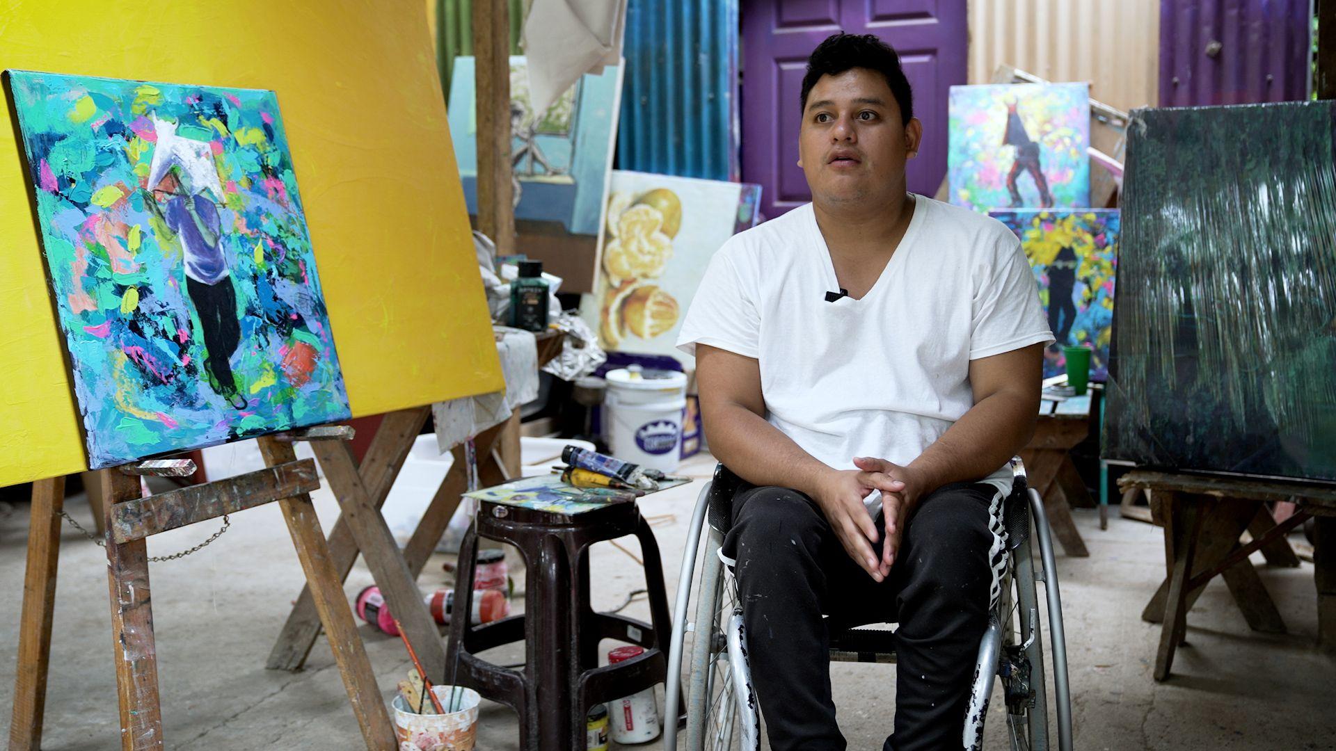 Un hombre en silla de ruedas junto a varios lienzos con pinturas de vivos colores. 