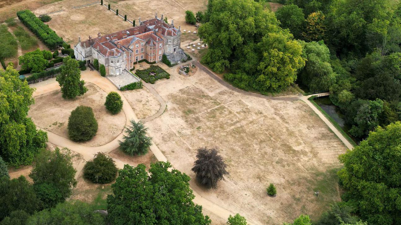 Una gran casa señorial está rodeada de hierba seca de color paja. Se pueden ver líneas geométricas tenues que parecen casas o edificios sobre la hierba desvaída