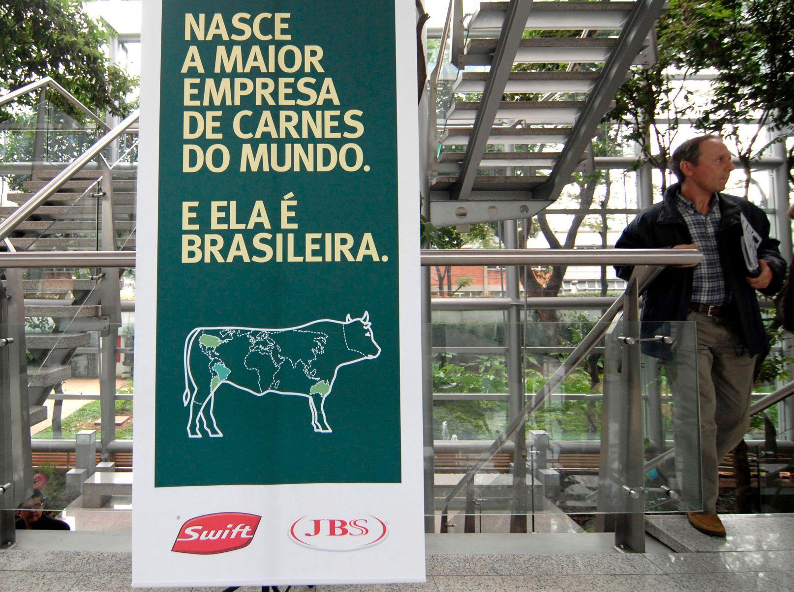 Cartel con la leyenda escrita en amarillo sobre fondo verde oscuro, con el dibujo de una vaca que tiene pintado el mapa mundi, y los logos de Swift y JBS abajo. 