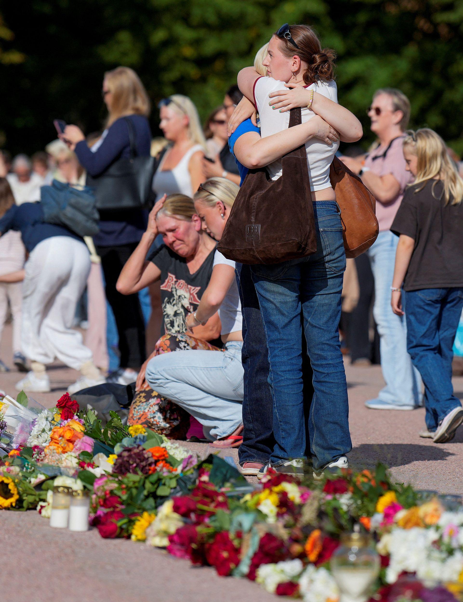 Dos personas se abrazan junto a una ofrenda floral al aire libre, mientras otras permanecen reunidas alrededor del homenaje.