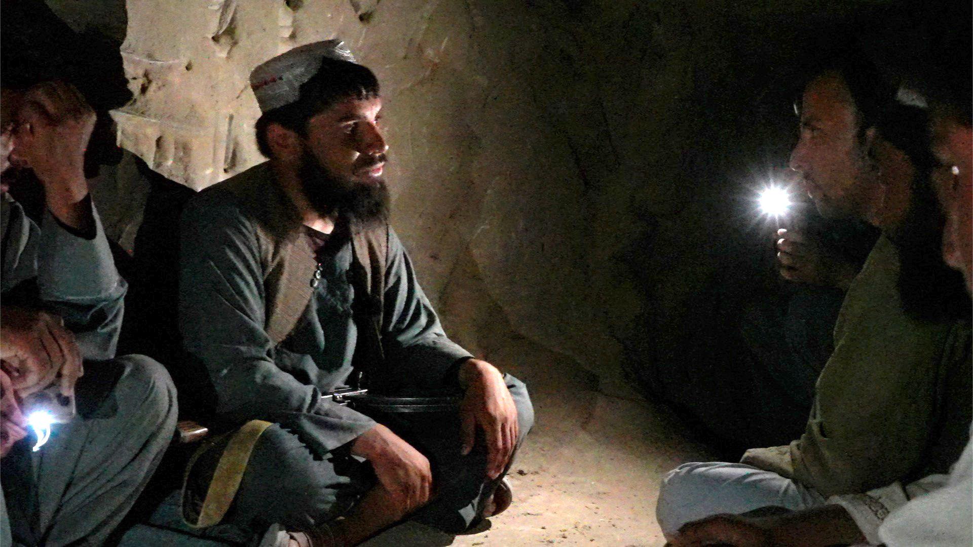 Un hombre barbudo, vestido con una túnica y pantalones oscuros tradicionales y un sombrero sin ala, está sentado con las piernas cruzadas en el suelo desnudo de una cueva oscura. Se encuentra a la izquierda de la imagen y mira a un hombre sentado a la derecha, que sostiene una antorcha. 
