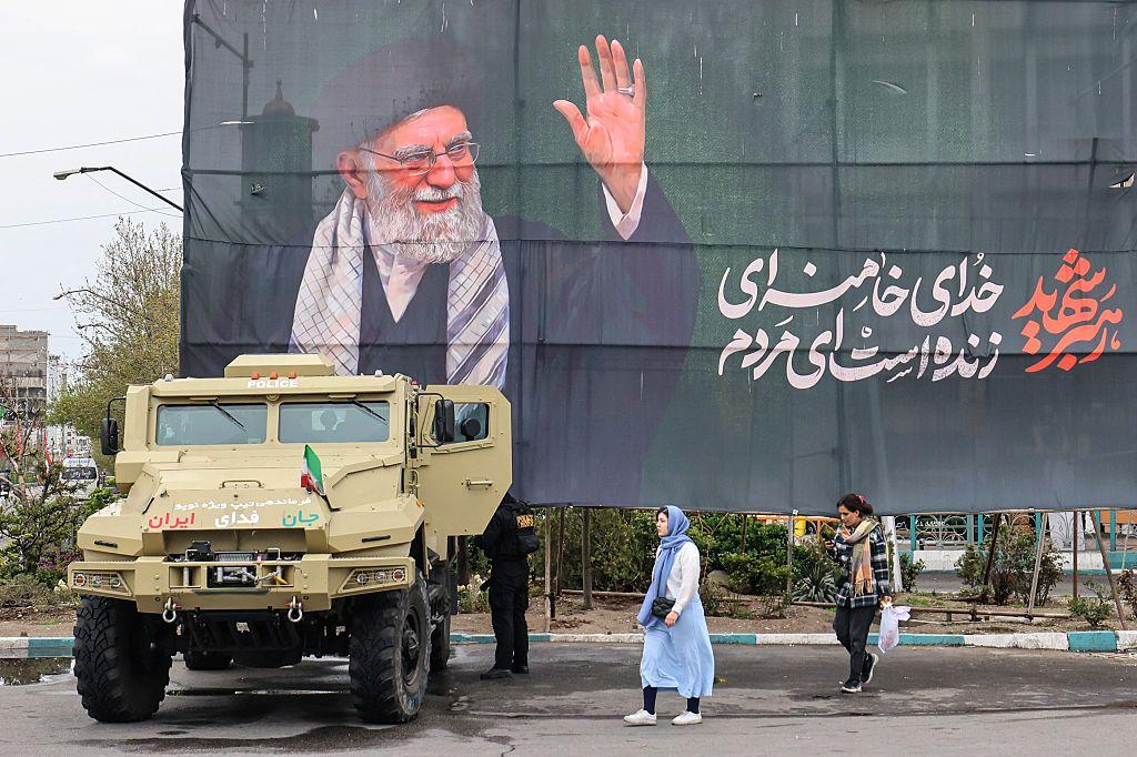 Mujeres pasan junto a un vehículo de las fuerzas de seguridad iraníes estacionado bajo una pancarta en honor al exlíder supremo de Irán.