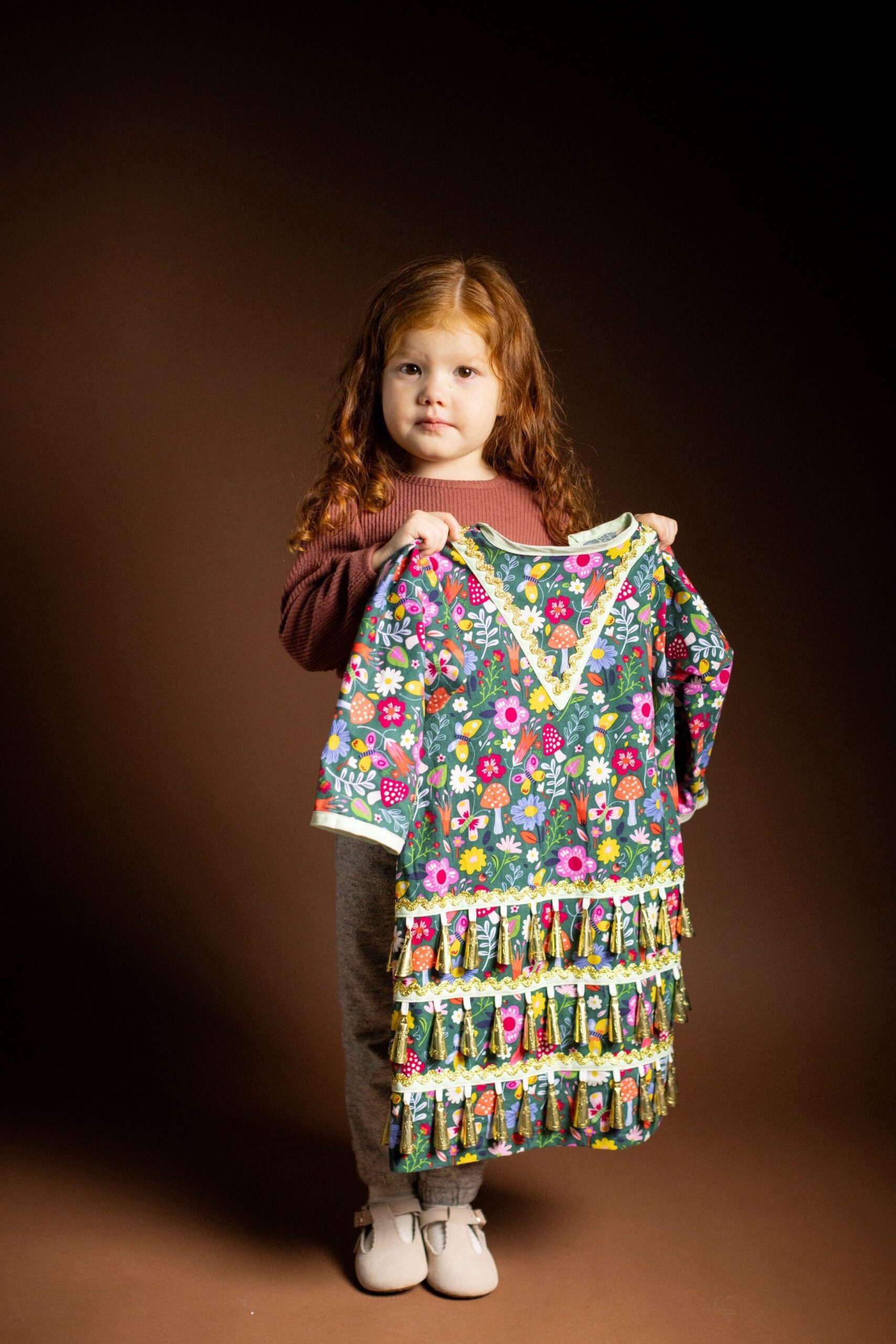 Retrato de estudio de cuerpo entero de una niña pequeña con cabello largo y rizado, de pie sobre un fondo marrón oscuro, sosteniendo un colorido vestido estampado con flores.