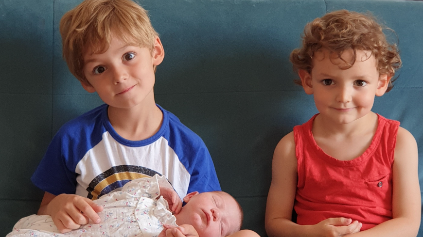 Dos niños pequeños, de entre tres y cinco años aproximadamente, están sentados en un sofá con un bebé recién nacido recostado sobre el regazo del mayor. Es un niño que mira a la cámara con una sonrisa (sin mostrar los dientes), con la cabeza ligeramente ladeada. La niña que está a su lado es menor. Existe un gran parecido familiar entre ambos, especialmente en sus ojos marrones.