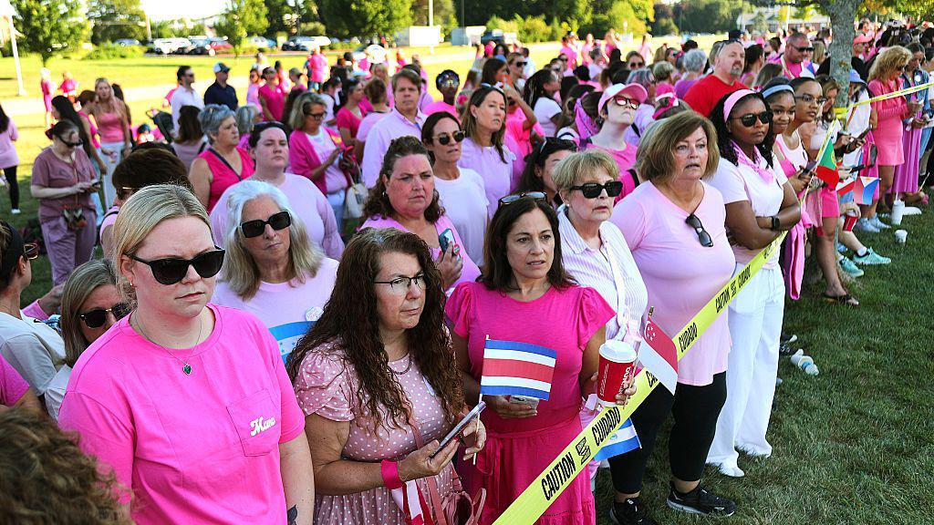 Mujeres con camisetas rosas se reúnen a las afueras del juicio de Clancy para apoyarla. Están de pie sobre el césped tras una cinta amarilla con el mensaje "Caution / Cuidado". Hay algunos hombres entre el grupo. 
