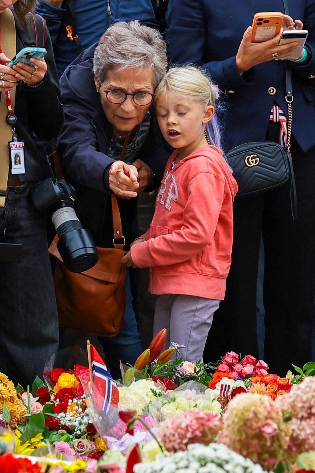 Una persona mayor señala una ofrenda floral con banderas de Noruega mientras acompaña a una niña; alrededor, varias personas observan el homenaje.
