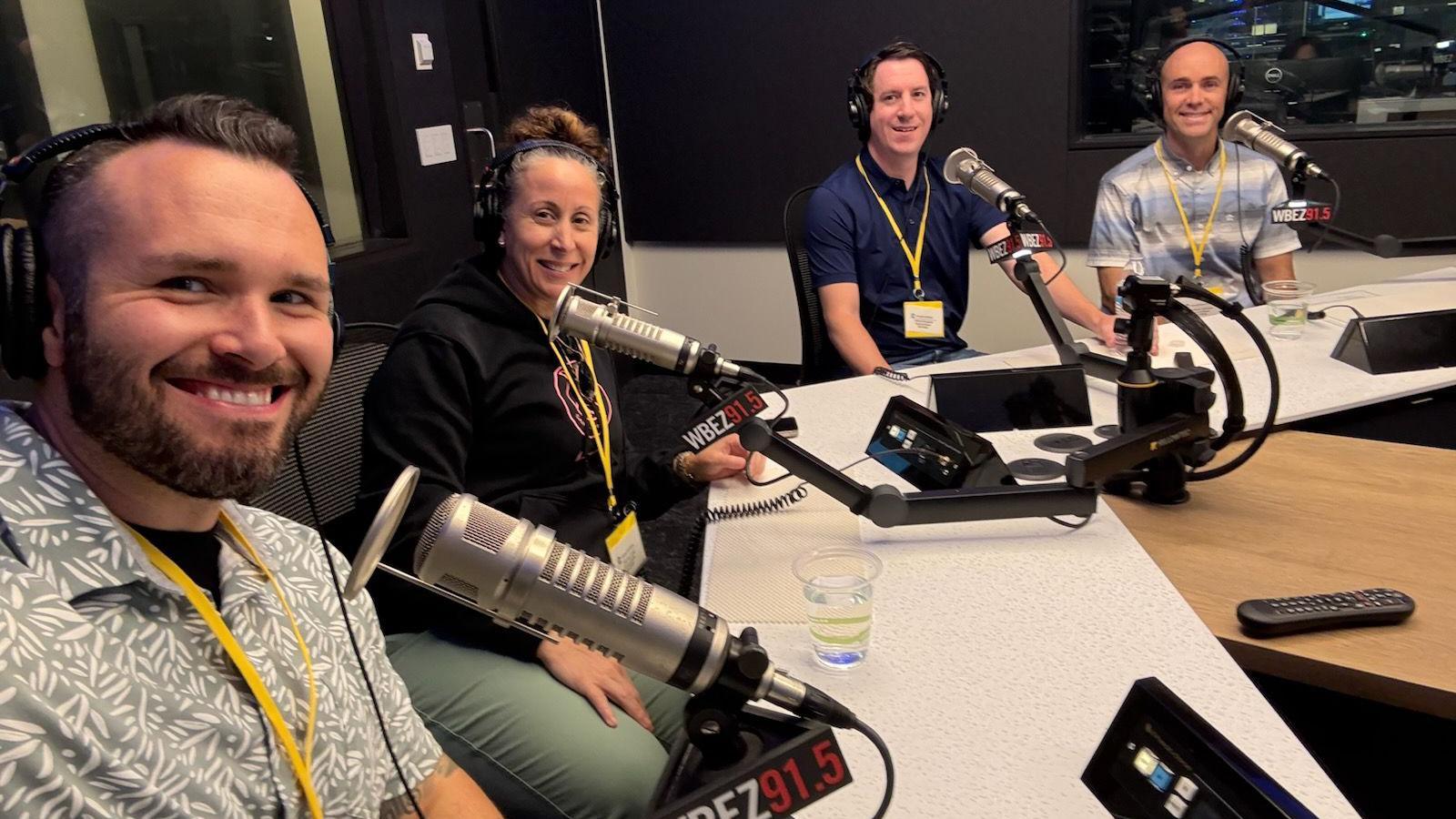 Tres hombres y una mujer, todos con auriculares puestos, están sentados en un estudio de grabación de podcasts con micrófonos frente a ellos.
