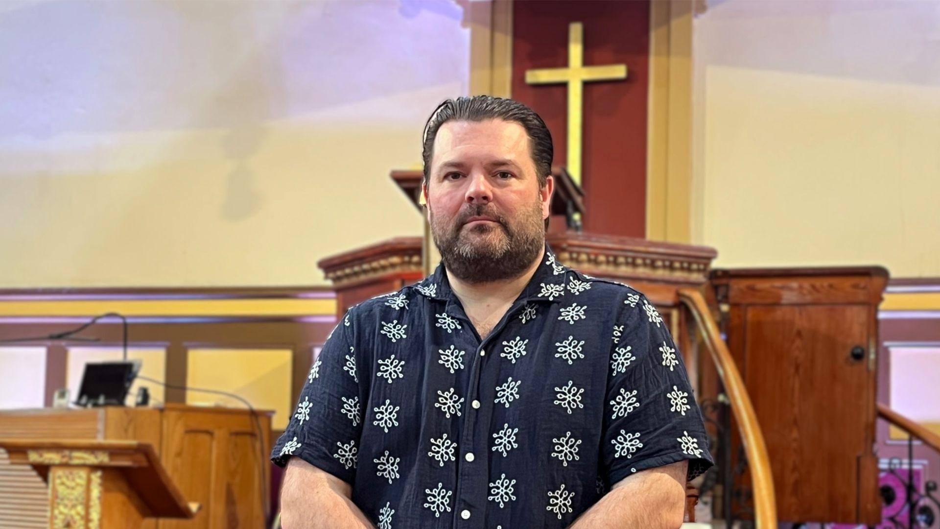 Jonathan se encuentra dentro de una iglesia, frente a un púlpito de madera con una cruz dorada sobre un fondo de madera.