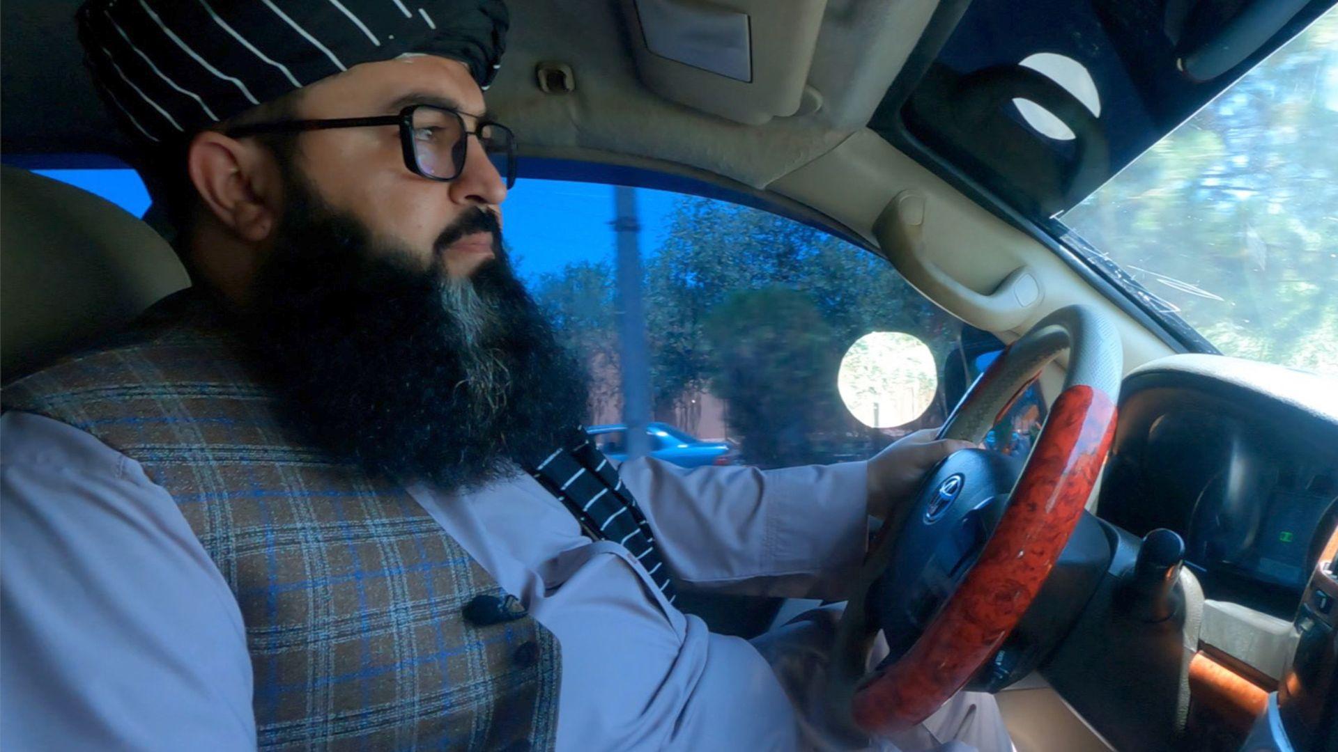 Vista lateral de un hombre conduciendo un coche, fotografiado desde el asiento del copiloto. Lleva barba poblada, gafas, un tocado negro con finas rayas blancas, una camiseta azul claro y un chaleco a cuadros.  