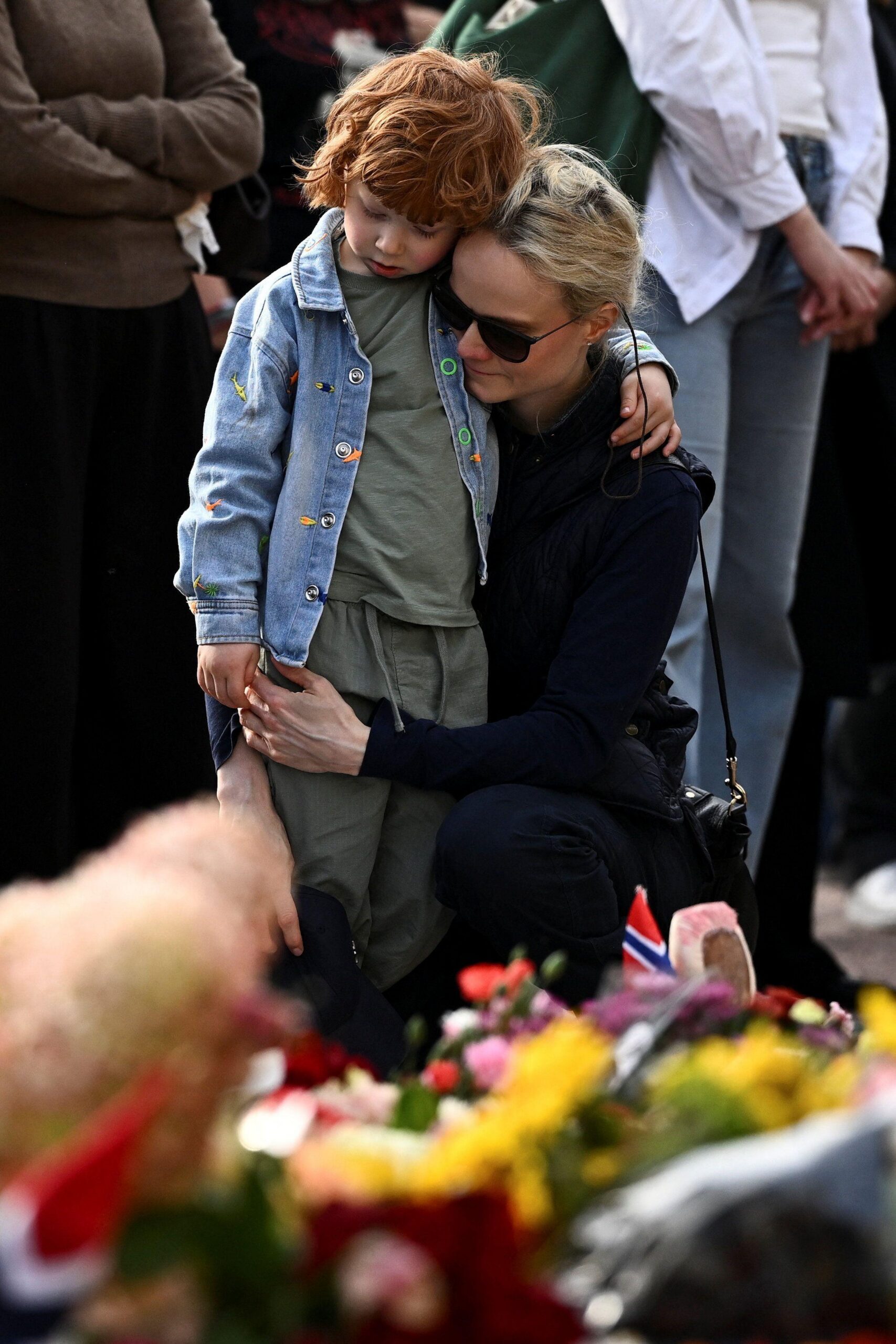 Mujer sostiene y abraza a un niño frente a una ofrenda floral con flores y una pequeña bandera de Noruega durante un homenaje público.