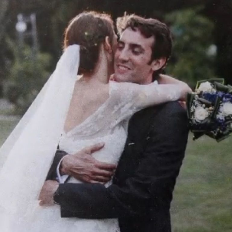 Una pareja casada se abraza en un jardín. La novia, de espaldas a la cámara y vestida con un vestido de novia blanco y un largo velo blanco, es alzada y abrazada por el novio, quien viste un traje oscuro. Ella sostiene un ramo de flores blancas y azules. 