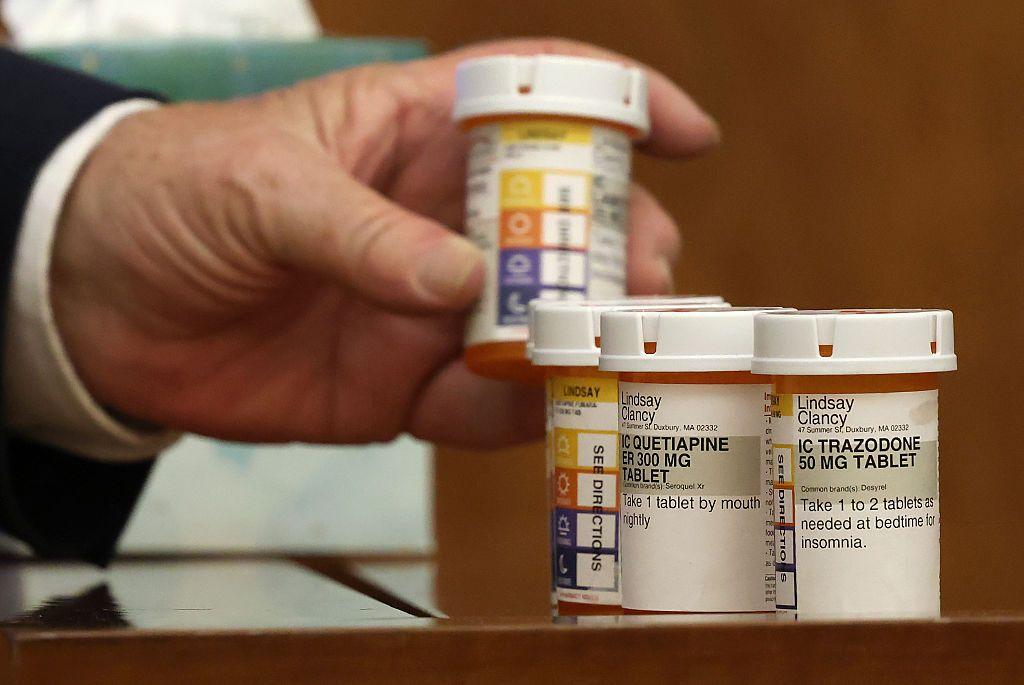 Una mano sostiene un frasco de medicamentos, otros dos están sobre una mesa.