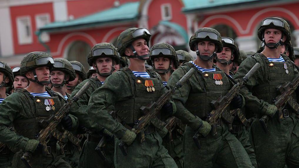 Un grupo de soldados armados con uniforme verde desfilan en el desfile del Día de la Victoria, que conmemora el fin de la Segunda Guerra Mundial, en Moscú, el 9 de mayo de 2026, con la pierna izquierda hacia adelante. El que está en el centro mira al frente con una amplia sonrisa, mientras que los demás miran a los lados, a la cámara, con expresión más seria.