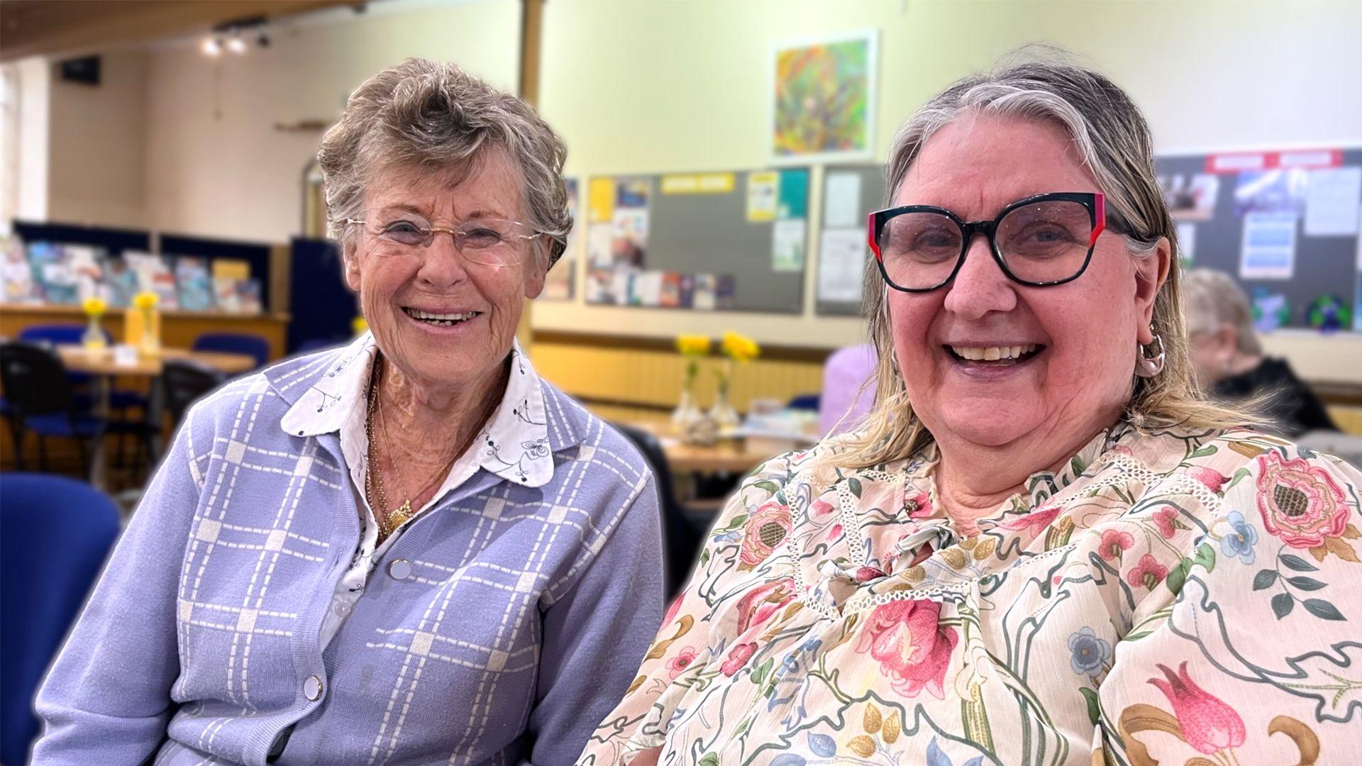 Muriel y Pamela, dos mujeres, están sentadas juntas en el interior de un espacio comunitario, sonriendo mientras conversan; ambas llevan gafas y blusas de colores vivos.