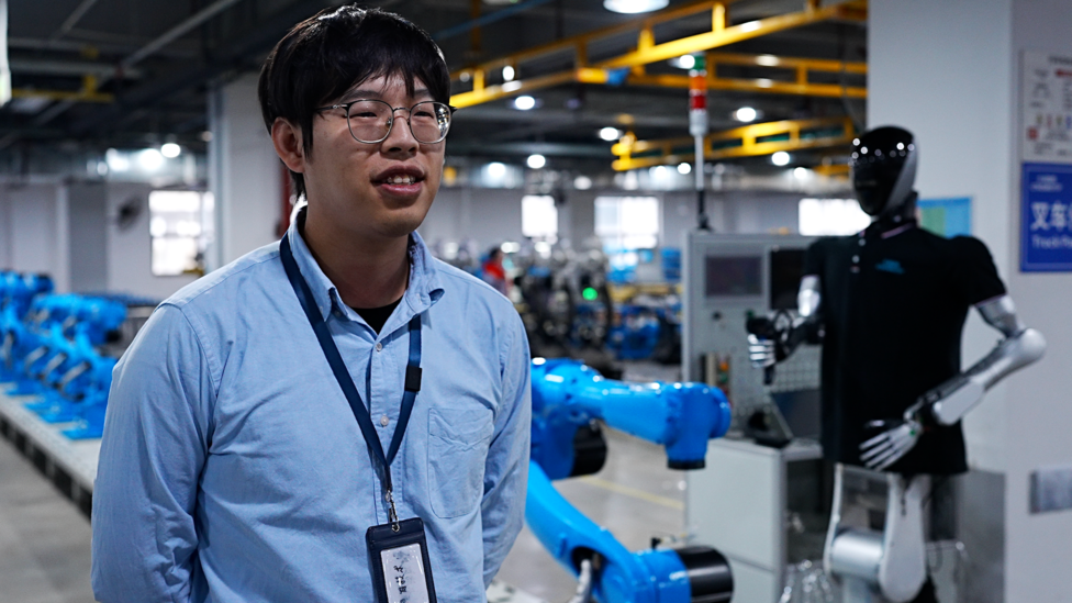 Hombre vestido con una camisa azul habla a la cámara mientras atrás hay un robot de color negro y una línea de producción robotizada de color azul. 