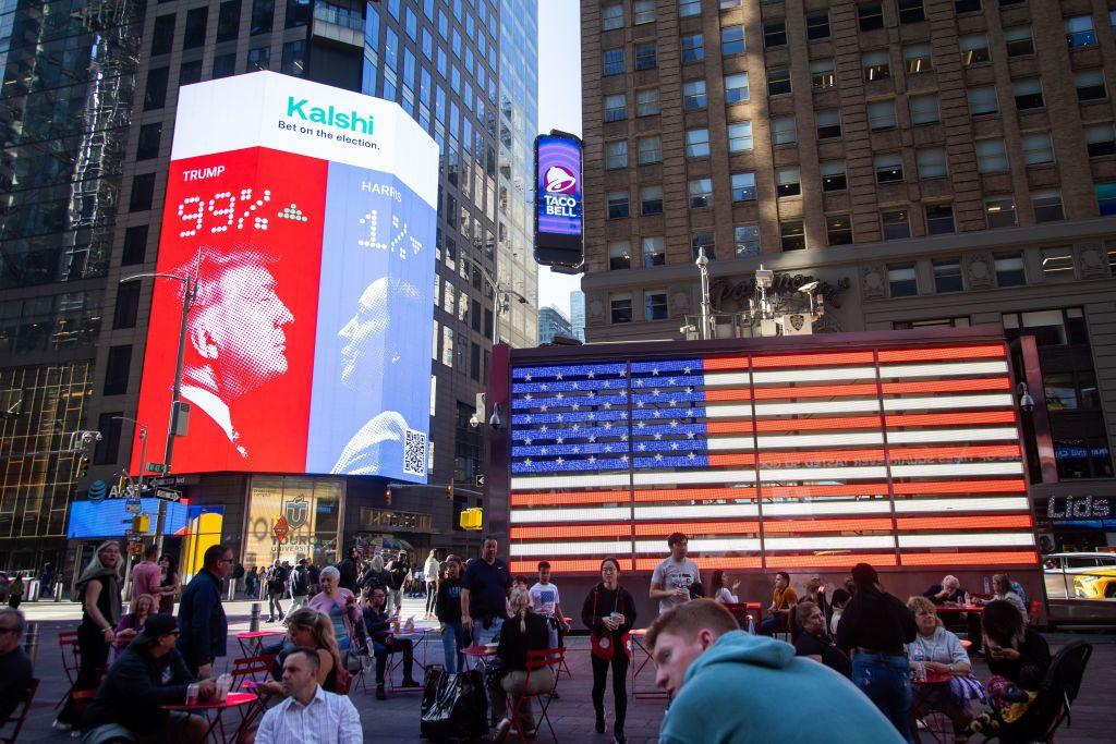 Edificios, gente y calle, y dos vallas publicitarias dominando: una con una bandera de EE.UU. enorme y la otra con los perfiles de los entonces candidatos Trump y Harris, y sus probabilidades de acuerdo a los usuarios de la app: 99% y 1% respectivamente. 