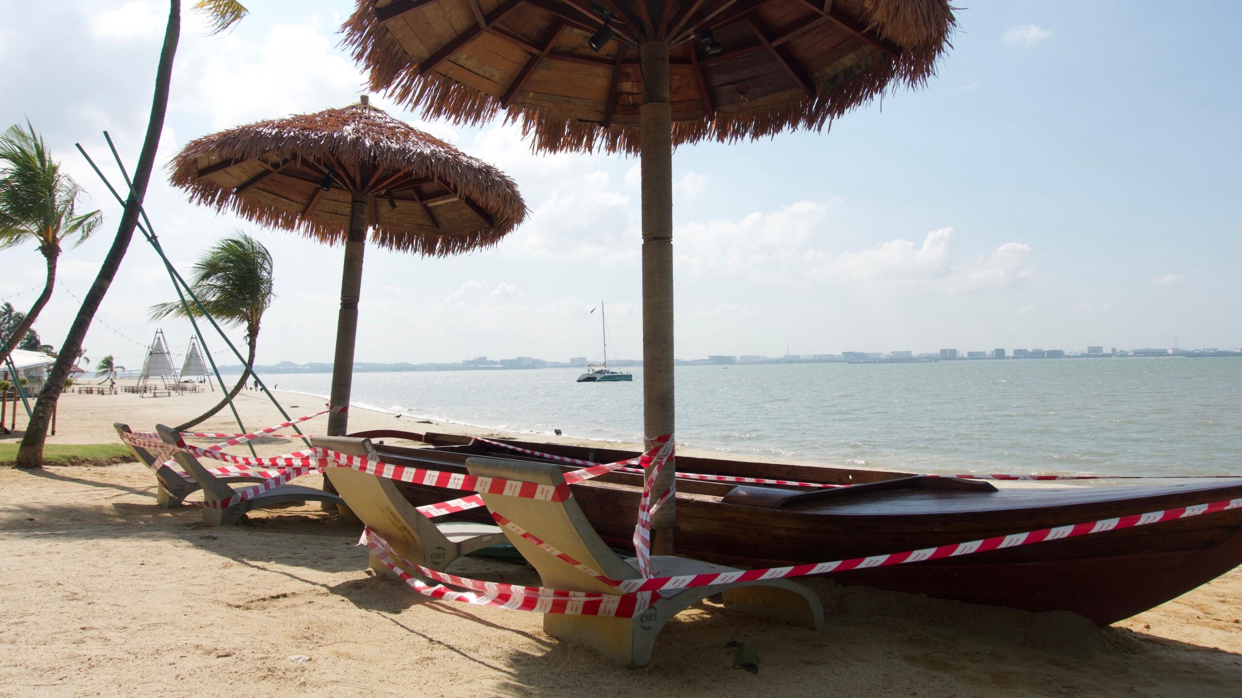 Un bote de madera convertido en banco en la playa, bajo sombrillas de hojas de palma y junto a sillas de piedra con forma de plátano, envuelto en cinta roja y blanca de "precaución". Al fondo se divisa el océano y, en el horizonte lejano, se vislumbra un tenue perfil de edificios.