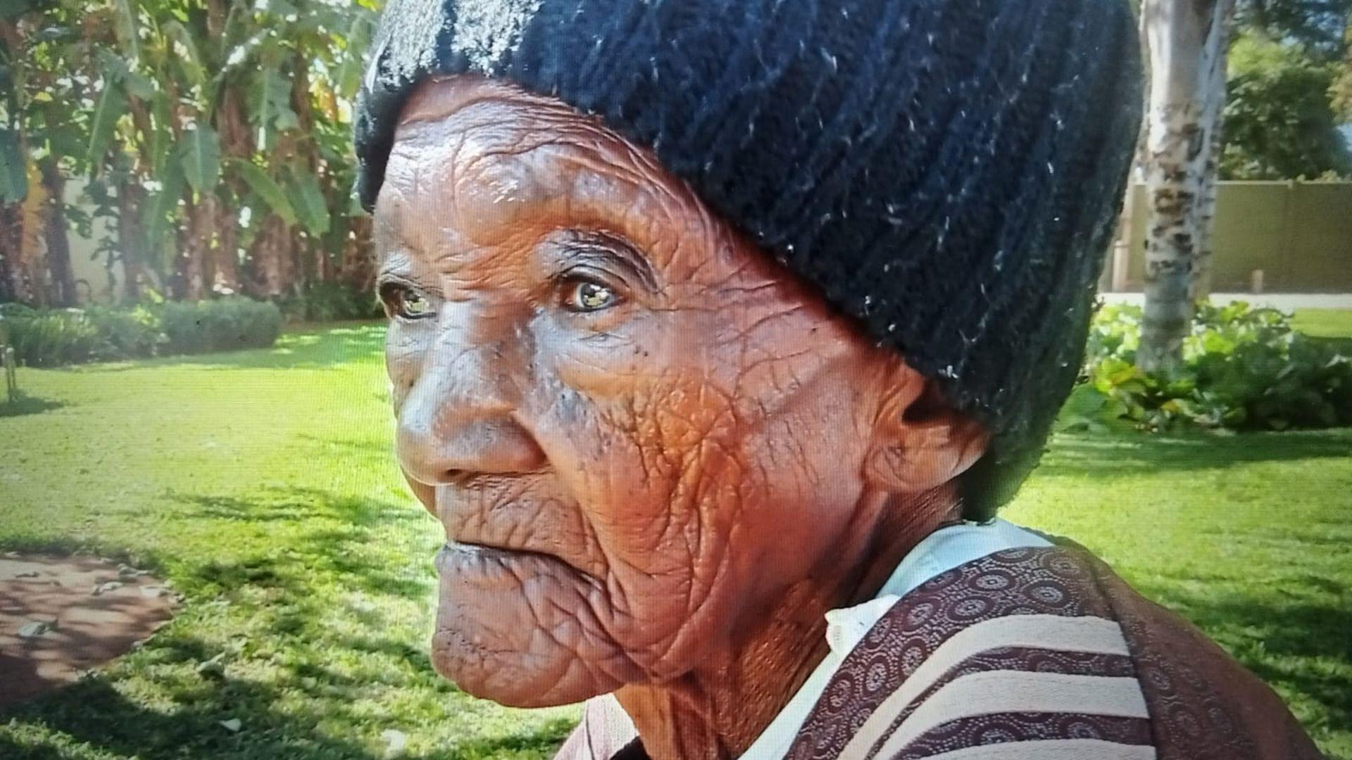 Una anciana vestida con un gorro de punto negro y un jersey marrón con estampados está sentada al aire libre en un jardín bañado por el sol. La imagen es de perfil en primer plano, resaltando profundas líneas faciales y una expresión pensativa mientras mira a lo lejos. Hierba verde exuberante, árboles y plantas frondosas llenan el fondo difuminado suavemente, creando un entorno natural y pacífico 