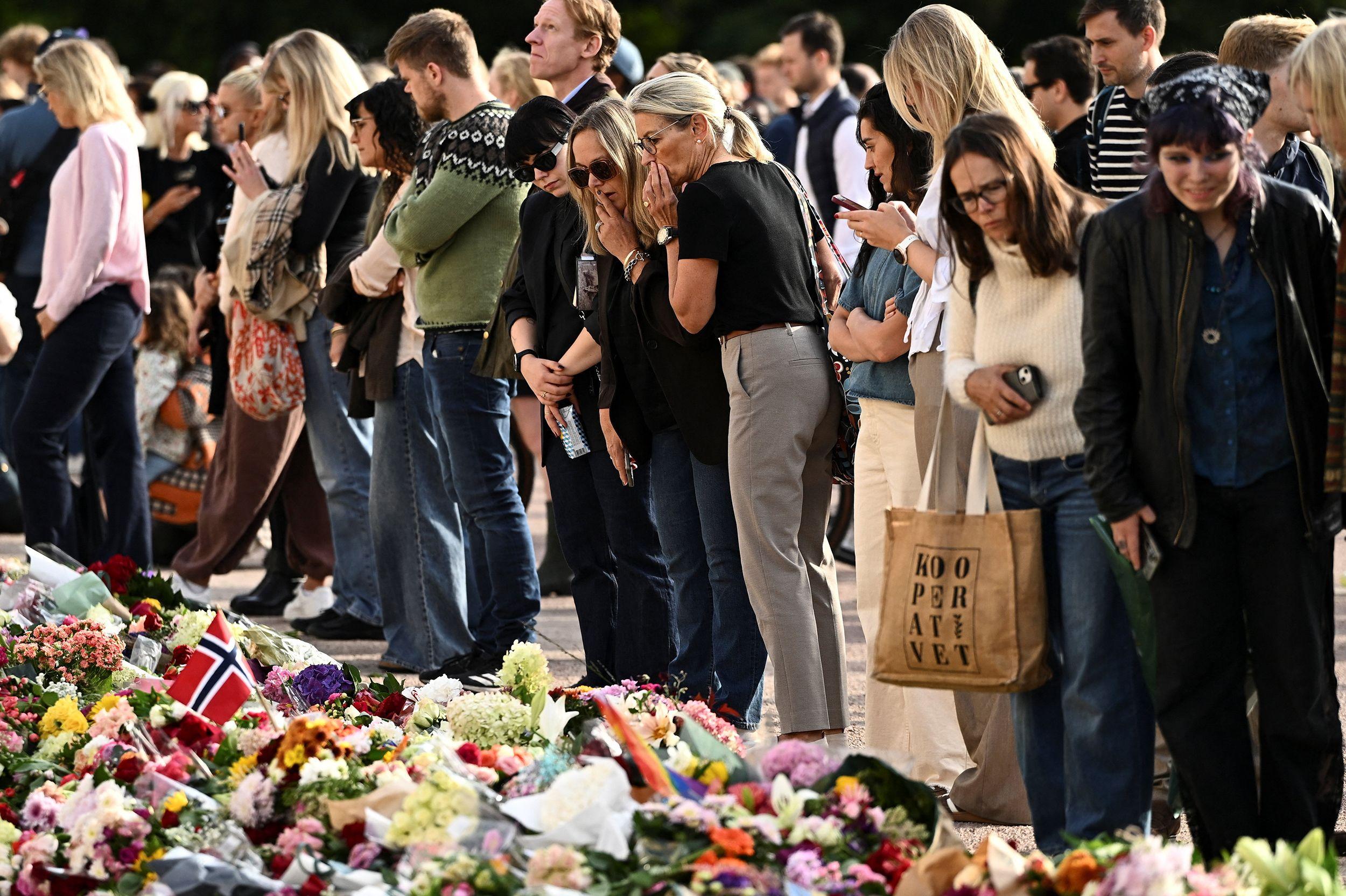 Personas observan una gran ofrenda floral con ramos y una bandera de Noruega colocados en el suelo durante una vigilia o homenaje público al aire libre.
