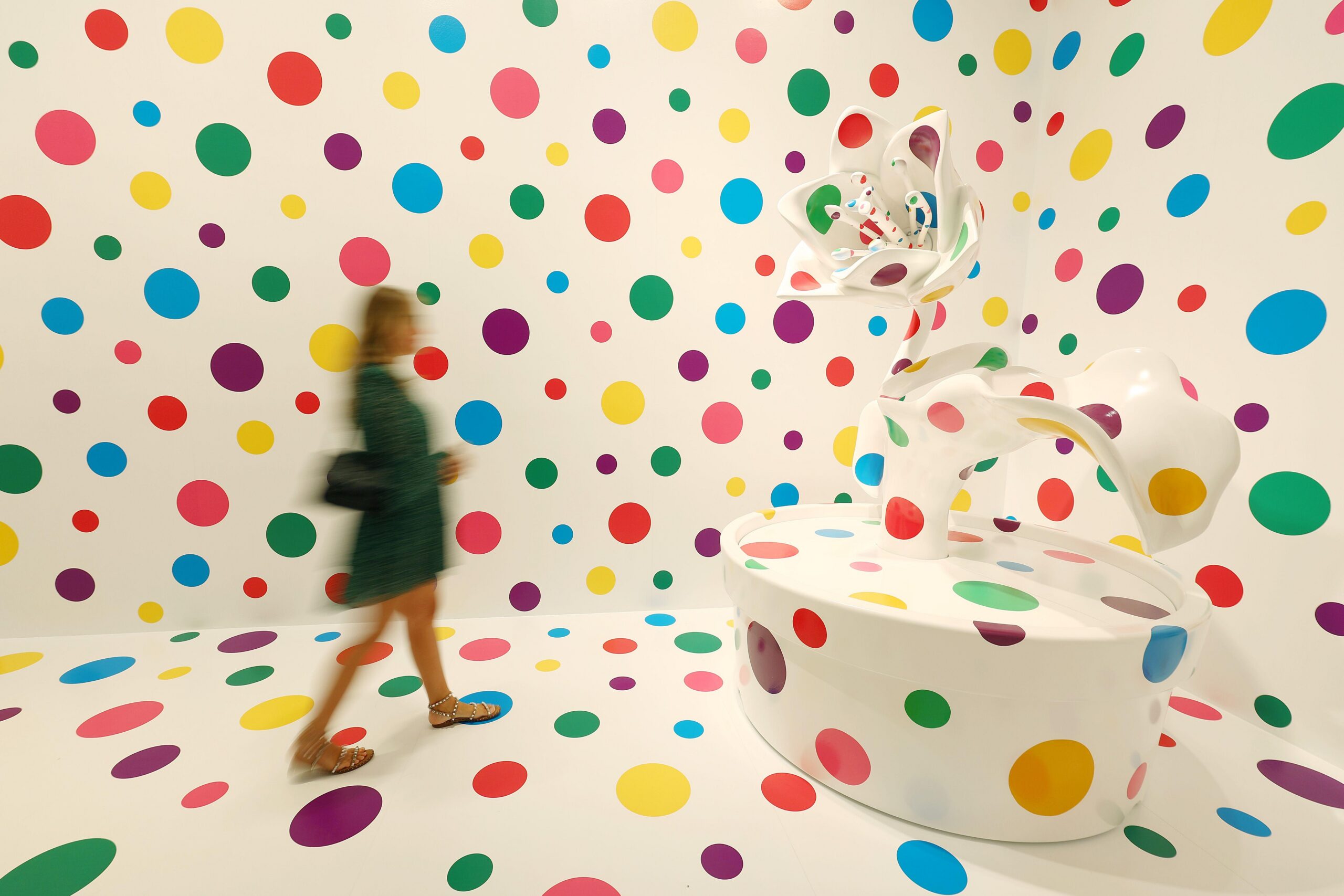 Un visitante de la galería, borroso, frente a una pared blanca y una escultura cubierta de manchas multicolores.