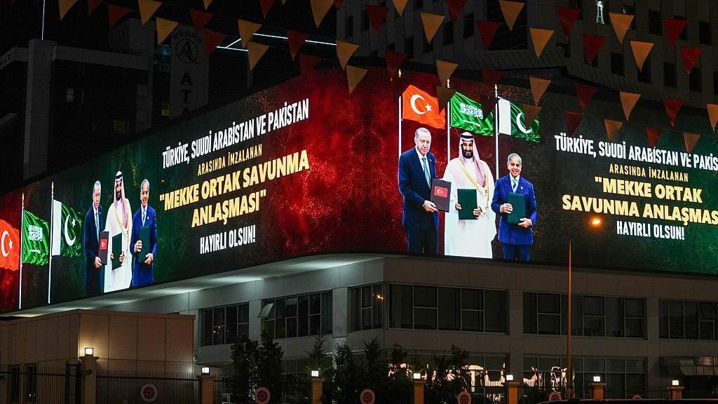 Fotografía tomada de noche. En una pantalla digital en lo alto del edificio, se ve a Erdogan, Bin Salman y Sharif sosteniendo documentos relacionados con el acuerdo. Detrás de ellos ondean las banderas de sus respectivos países. Junto a los líderes, en letras grandes, se lee: "Que el Acuerdo Conjunto de Defensa de La Meca, firmado entre Turquía, Arabia Saudita y Pakistán, sea propicio".