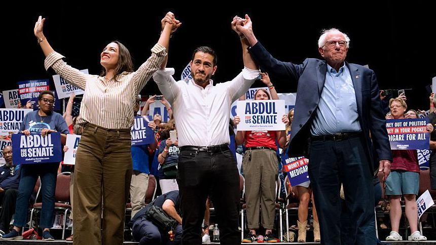 Alexandria Ocasio-Cortez, Abdul El-Sayed y Bernie Sanders están tomados de las manos y las levantan durante un evento de campaña en el que varias personas levantan carteles de Abdul para el Senado.