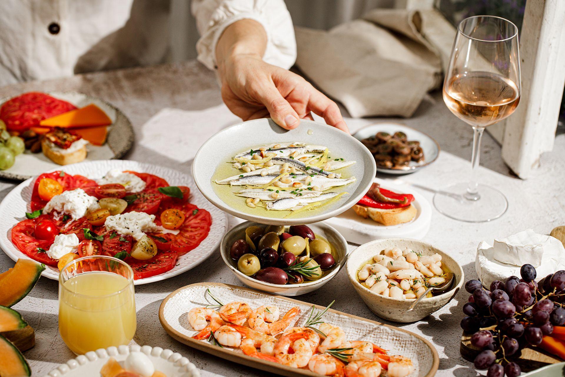Una mano sirve un plato de boquerones marinados sobre una mesa llena de tapas mediterráneas, que incluyen gambas, aceitunas, tomates, queso, judías, uvas, melón y una copa de vino blanco.