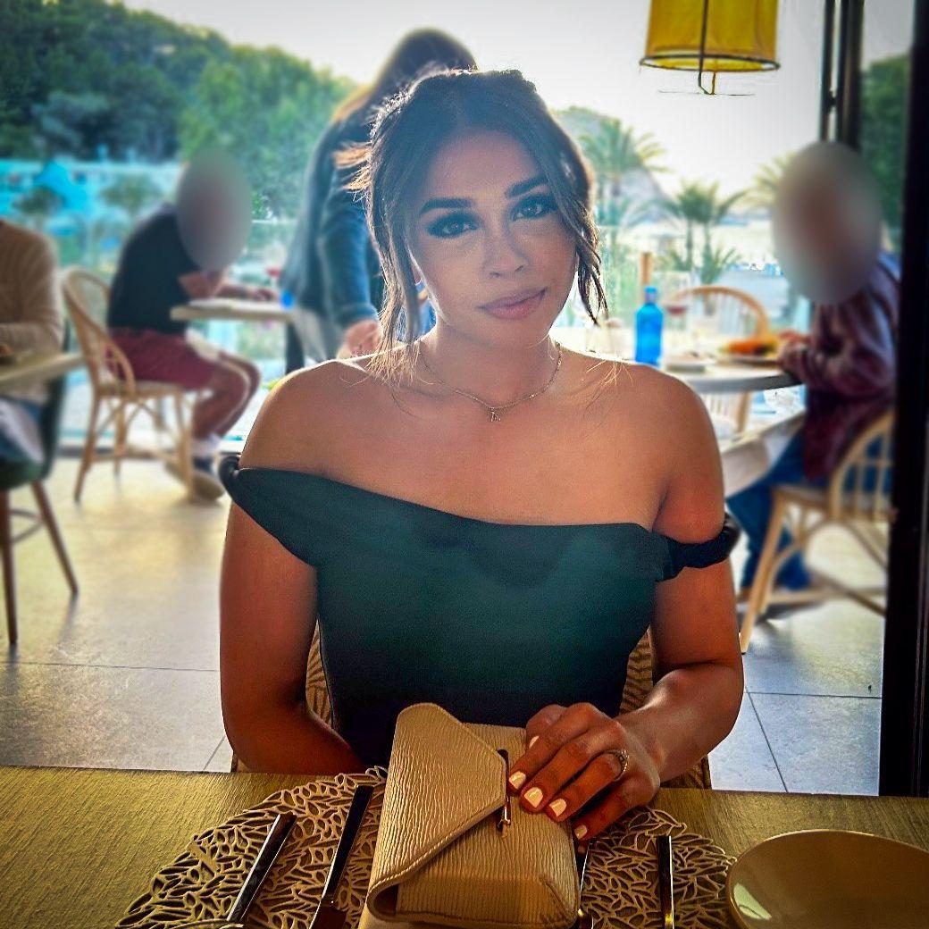Una foto de una joven sentada en una mesa en un restaurante concurrido. Su pelo castaño está recogido hacia atrás y lleva una camiseta negra descubierta con hombros descubiertos, con su bolso sobre la mesa