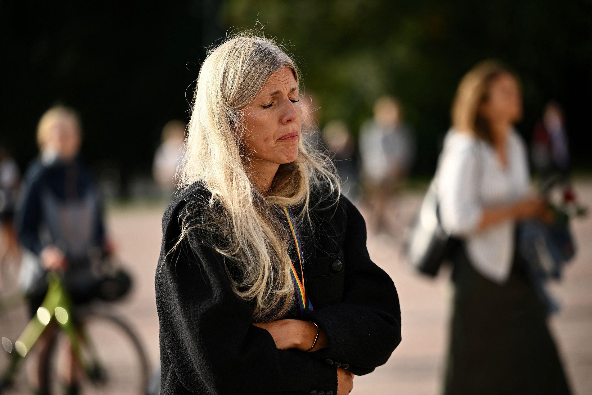 Mujer de cabello largo y abrigo oscuro permanece de pie con los brazos cruzados durante una concentración al aire libre, con asistentes desenfocados al fondo.
