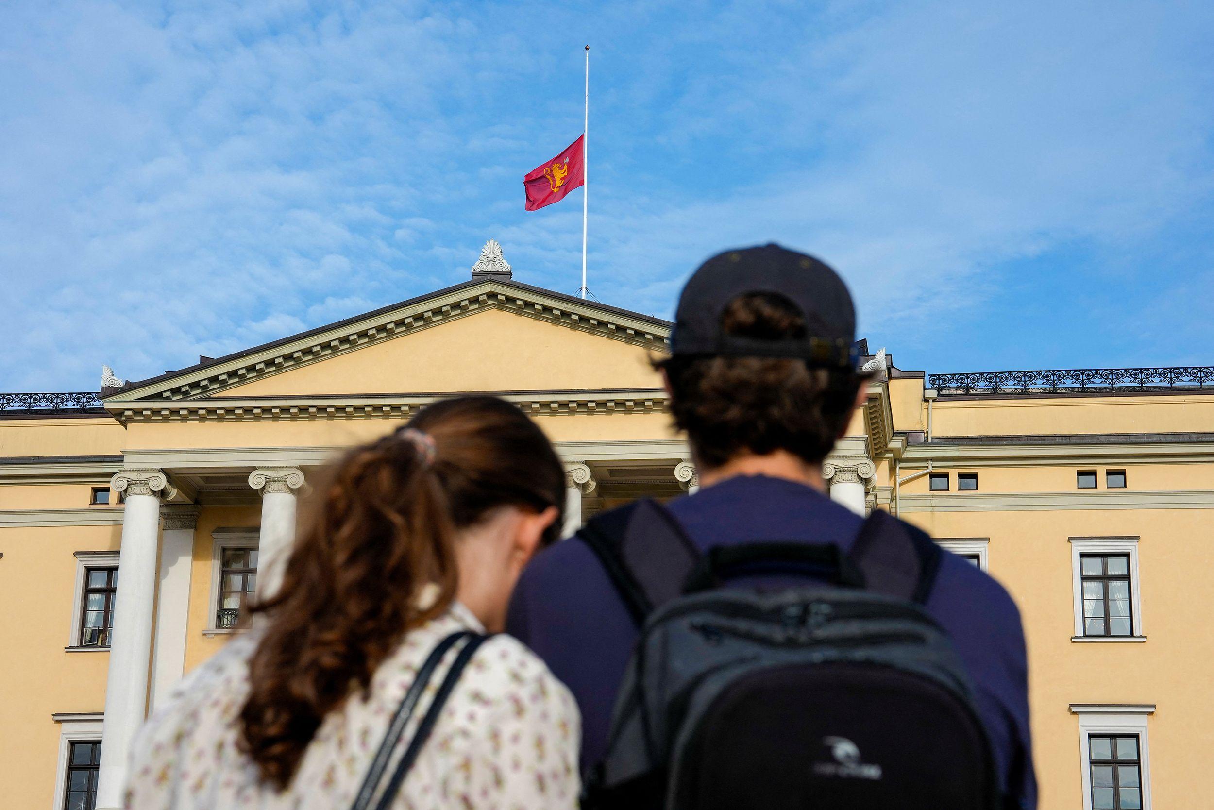 Dos jóvenes observan un edificio oficial mientras una bandera ondea a media asta en el techo bajo un cielo parcialmente nublado.
