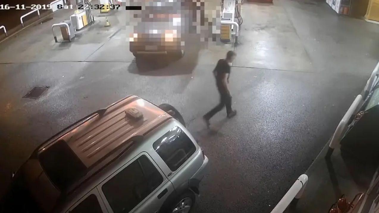 Una imagen de cámara de seguridad muestra a una persona caminando en una gasolinera durante la noche; se observan varios vehículos estacionados y una marca de tiempo visible en la esquina superior izquierda.
