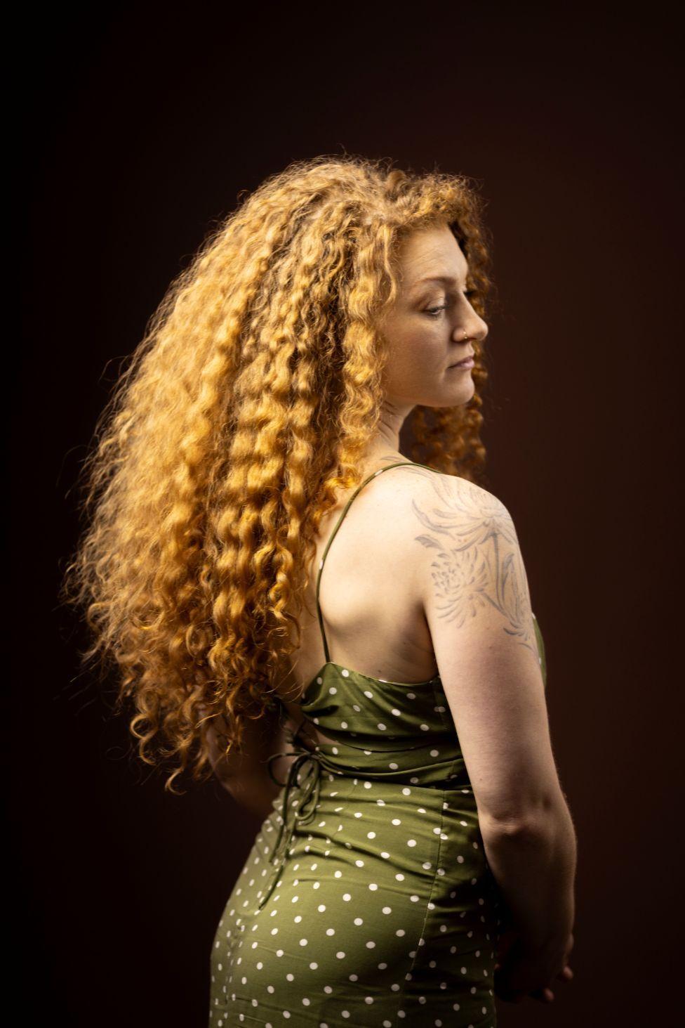 Retrato de estudio de una persona con cabello largo, voluminoso y rizado, fotografiada de espaldas y de perfil, girando ligeramente hacia la cámara. 