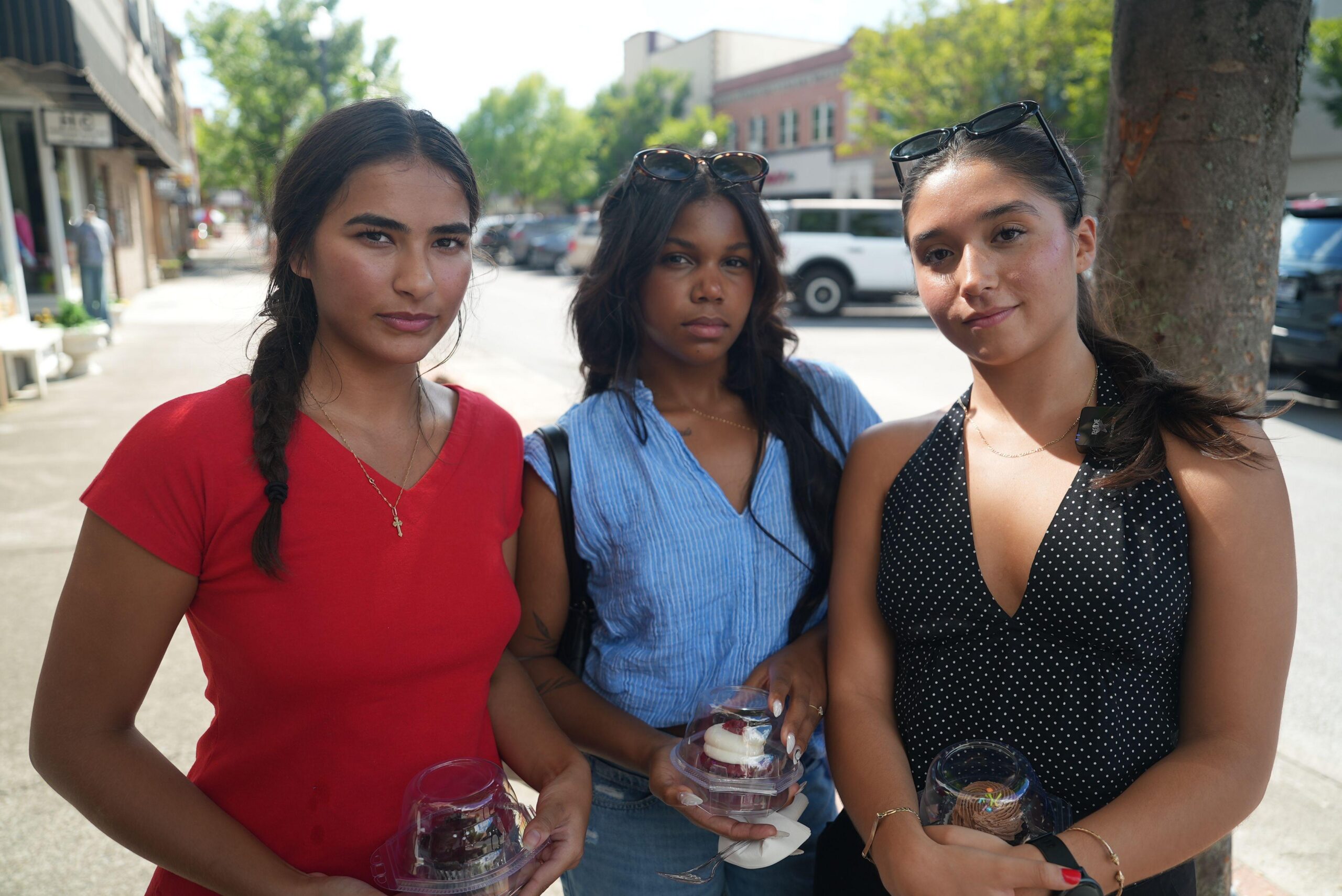 Tres mujeres de 21 años están de pie sosteniendo pasteles recién comprados en una tienda de una de las principales calles de Kingsport en el centro. Una lleva una camiseta roja de escote en V, un collar con cruz y su largo pelo oscuro trenzado. La amiga de al lado lleva gafas de sol en la cabeza y una camisa azul de escote en V por encima de los vaqueros. La tercera, con el pelo recogido en una coleta, lleva una camiseta negra tipo halter y gafas de sol en la cabeza