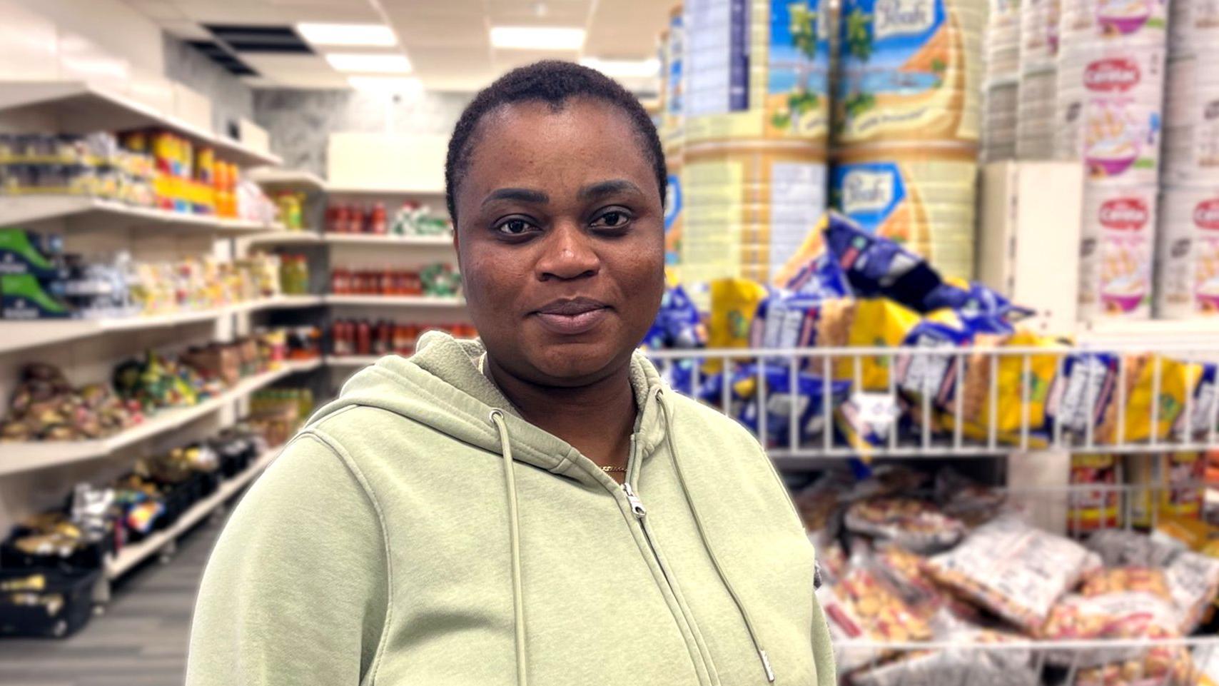 Christianah lleva una sudadera con capucha de color verde claro y está de pie dentro de un supermercado bien iluminado, con estantes llenos de productos enlatados, aperitivos y alimentos a granel detrás de ella.