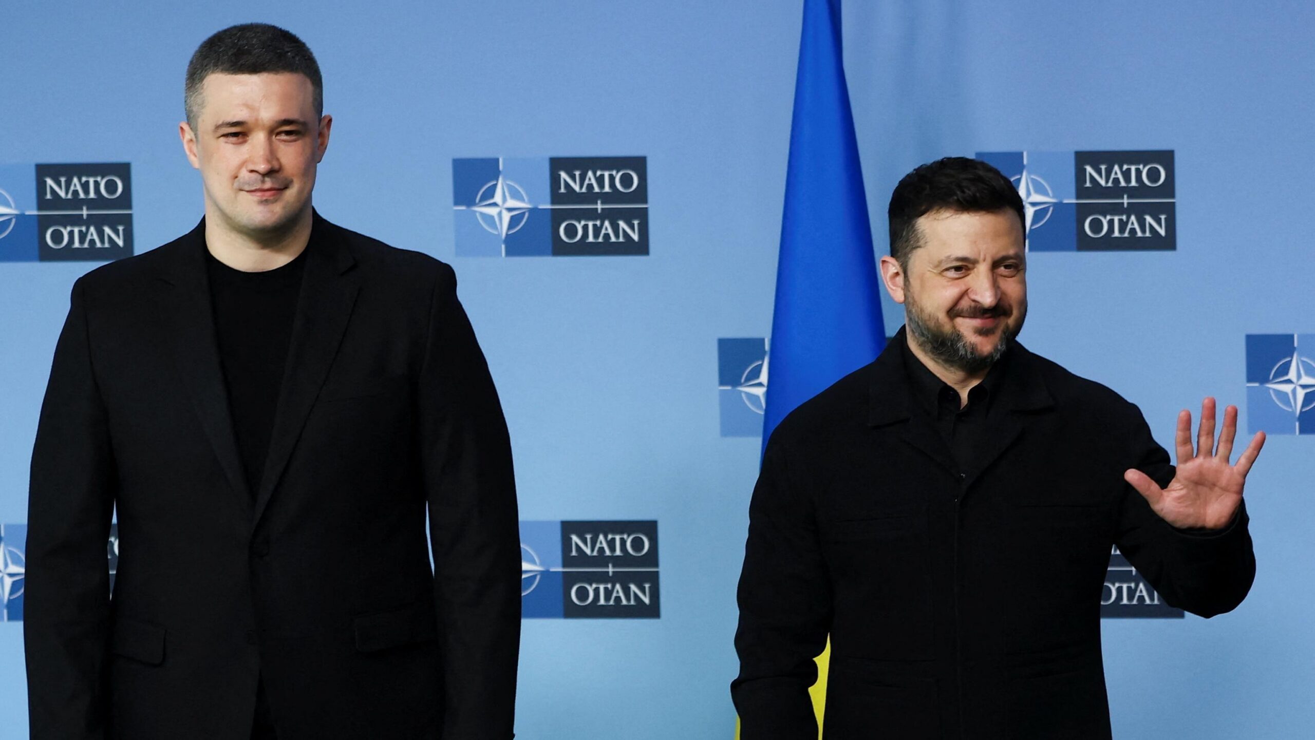 El presidente Volodymyr Zelensky aparece de pie, sonriendo y chocando las manos con otro político, también vestido de negro, frente a un fondo azul con el símbolo de la OTAN y la bandera ucraniana.