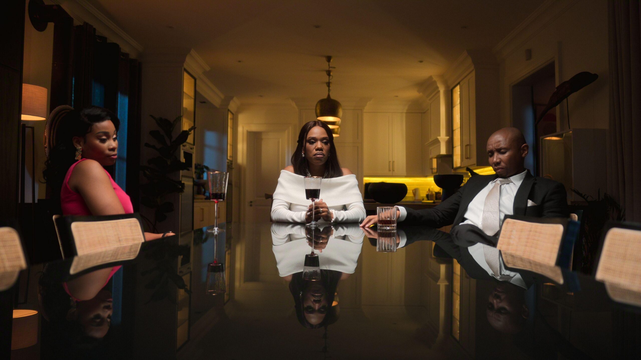 Una tensa escena de la serie de Netflix. Reflejados en una mesa negra y brillante está una mujer con un vestido rosa sentada a la izquierda, una mujer con un vestido blanco de hombros descubiertos sostiene una copa de vino en el centro, y un hombre con esmoquin negro sentado a la derecha, todos con expresiones serias y abatidas en un ambiente tenue,  dentro de un comedor de alta gama