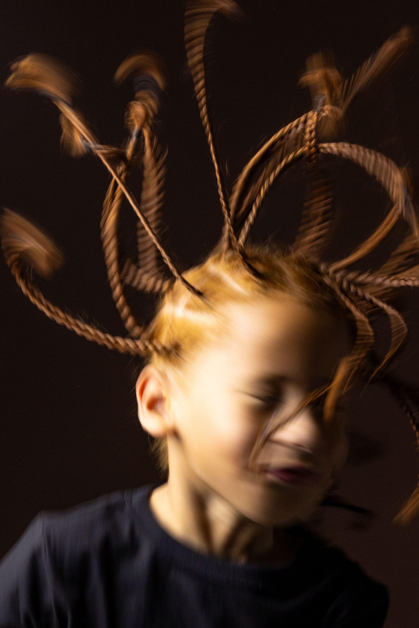 Retrato artístico en primer plano de una niña con múltiples trenzas que se extienden hacia afuera en movimiento, creando un dinámico efecto borroso. 